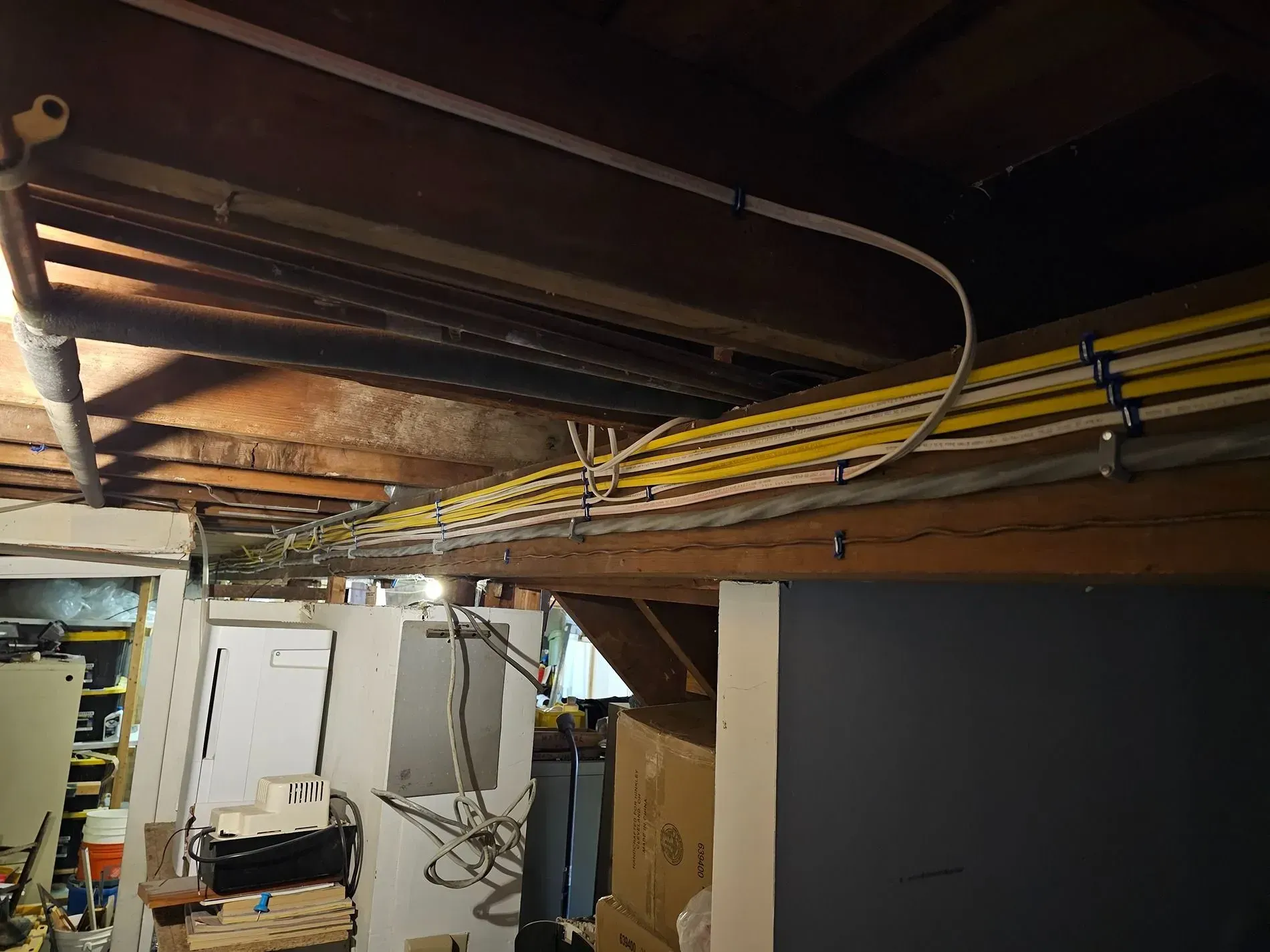 Basement ceiling with exposed joists, yellow pipes, and wiring above storage and utility equipment