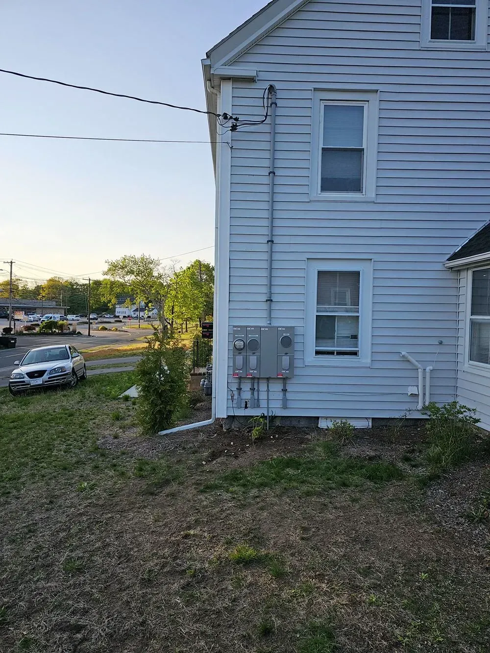 Side of a light-blue house with utility meters, overlooking a grassy yard and street at sunset.