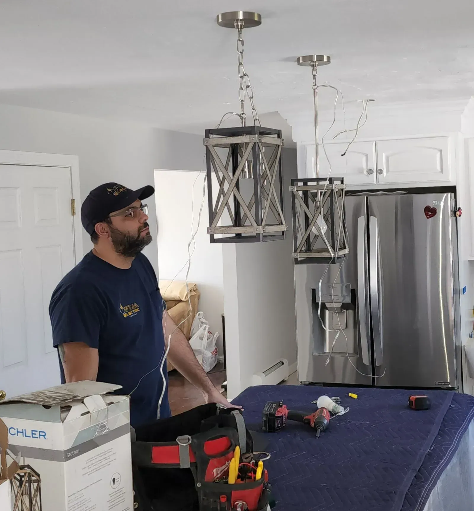 Man standing in a kitchen beside a table of tools, with hanging light fixtures and a stainless-steel refrigerator