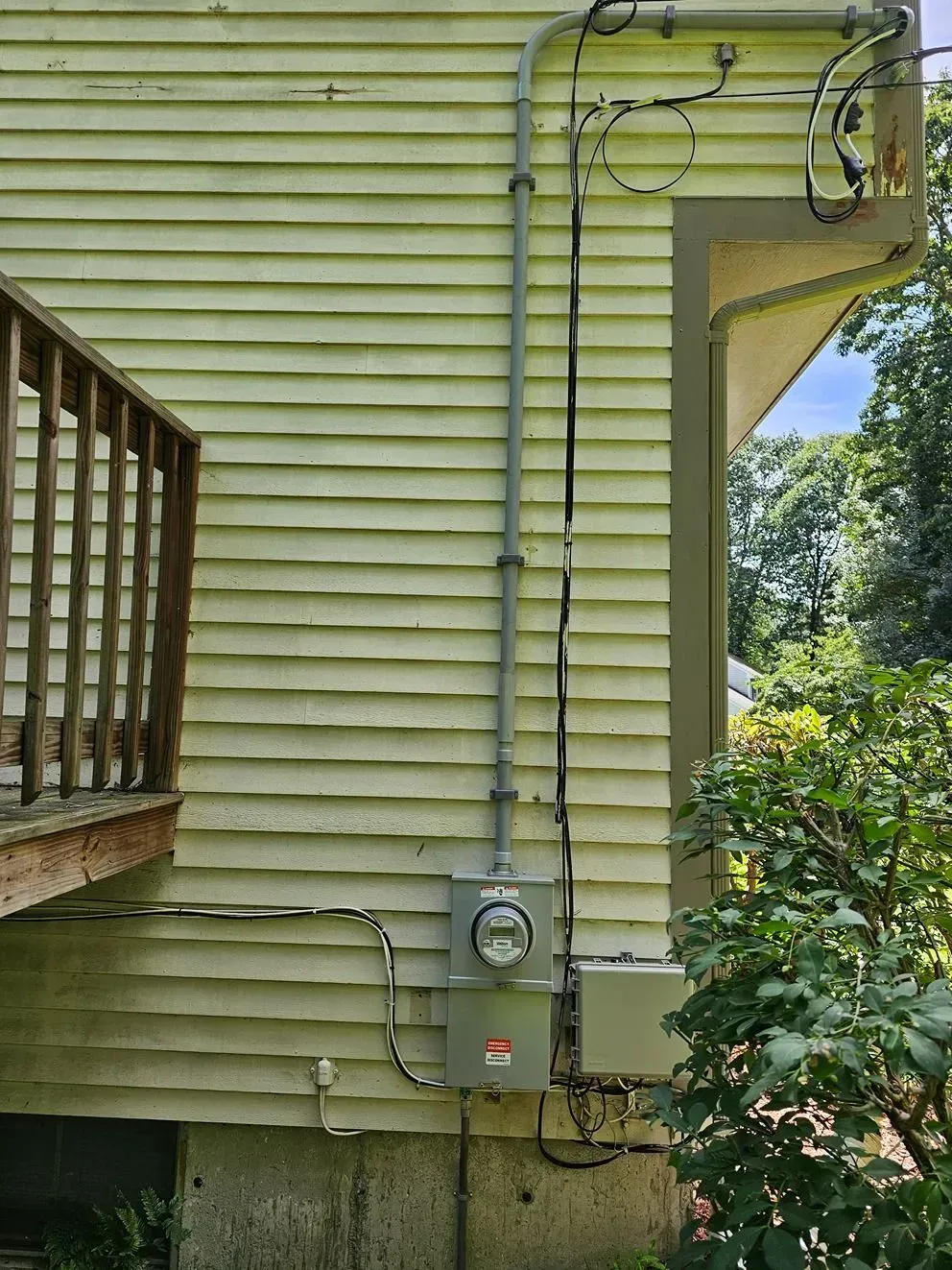 Yellow house exterior with utility meter, conduit, and overhead wires beside a porch and bushes