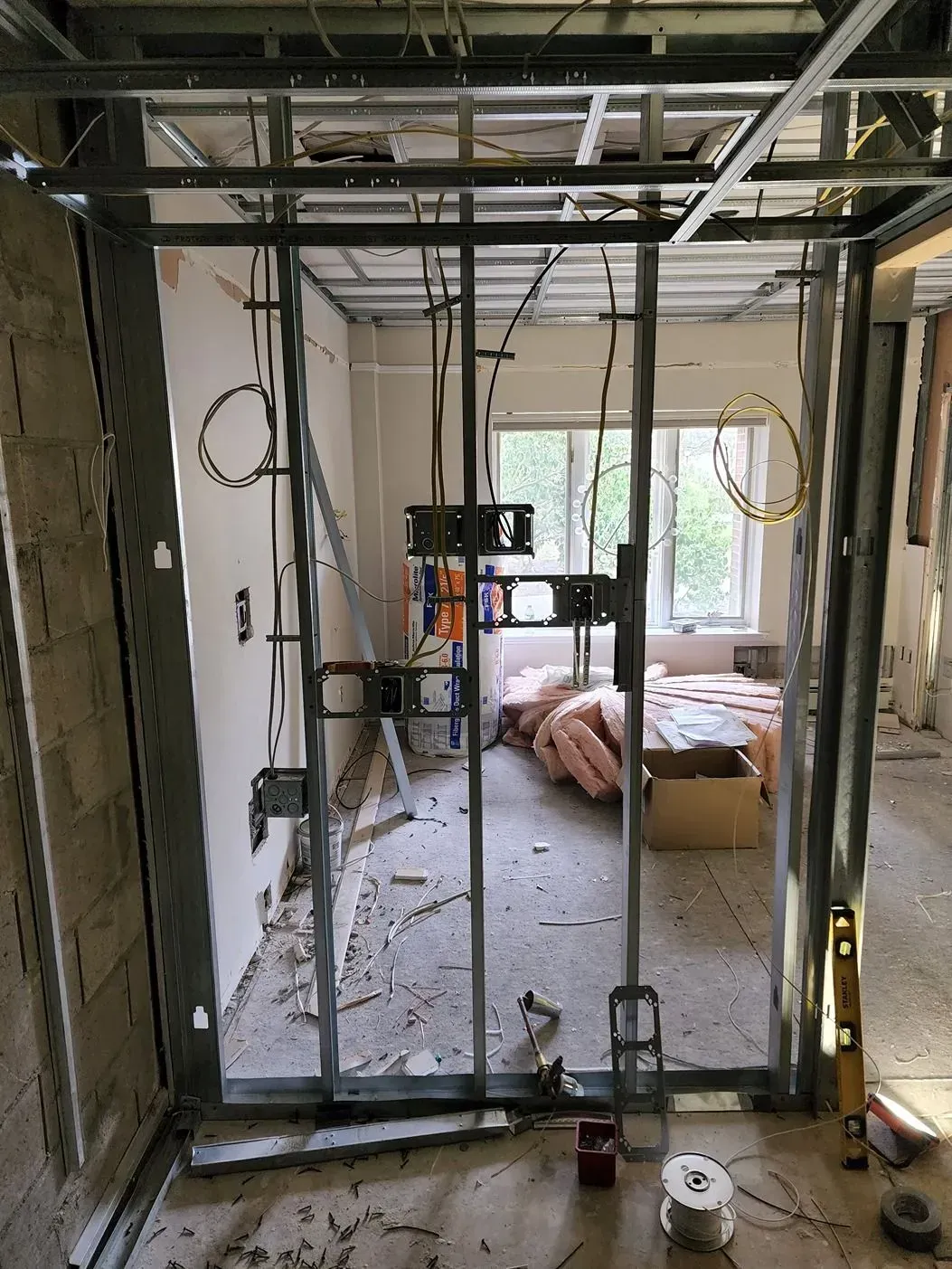 Partially framed room under construction with exposed studs, hanging wires, and a window overlooking a bed.