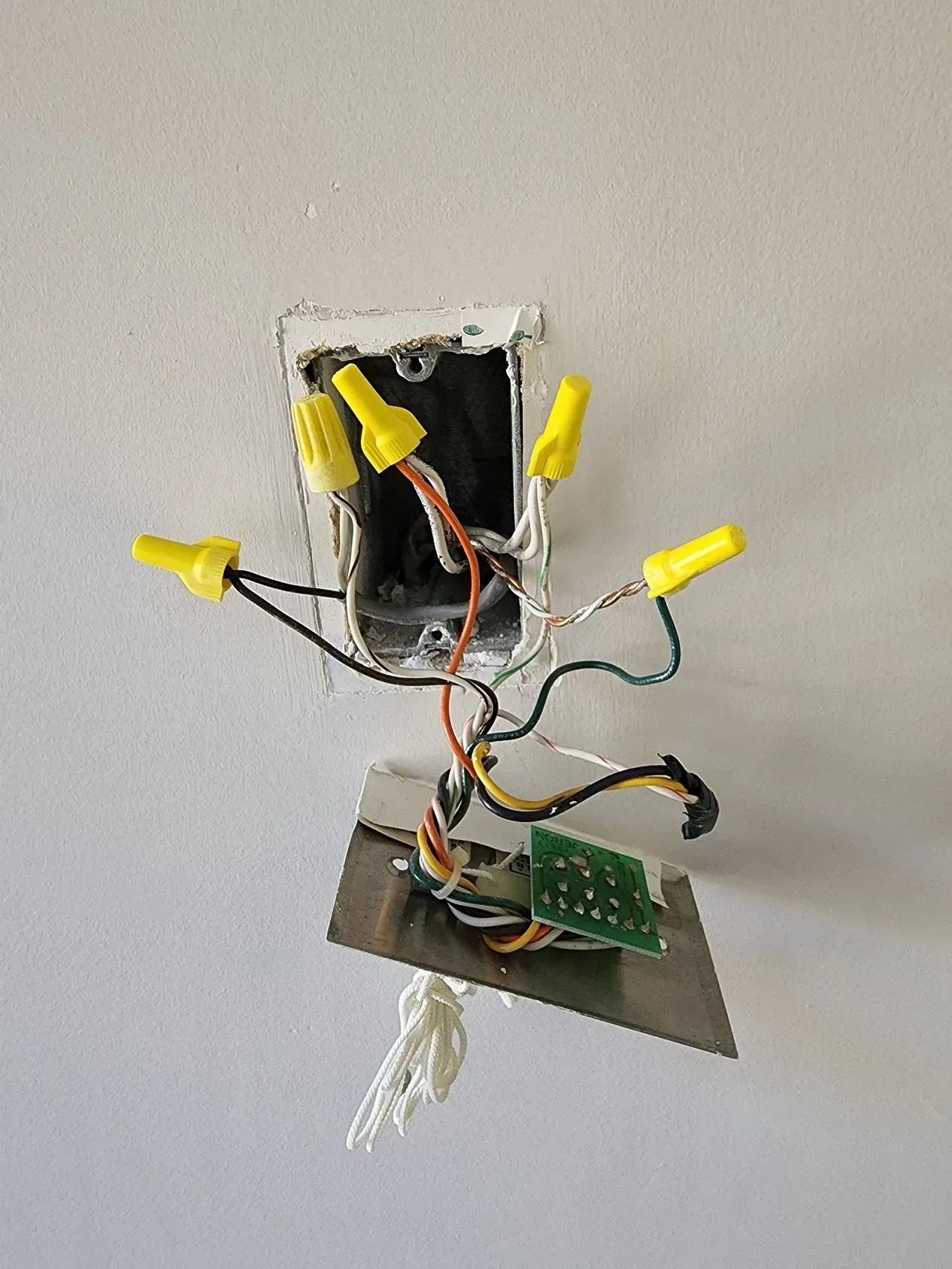 Exposed electrical wiring with yellow wire nuts and a hanging circuit board in a wall opening