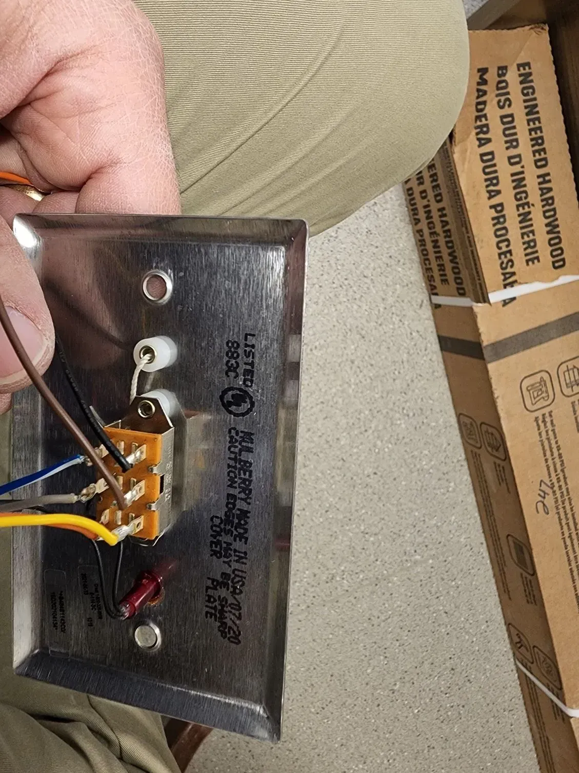 Hand holding a small wired electronic device with metal backplate and attached orange and white wires.