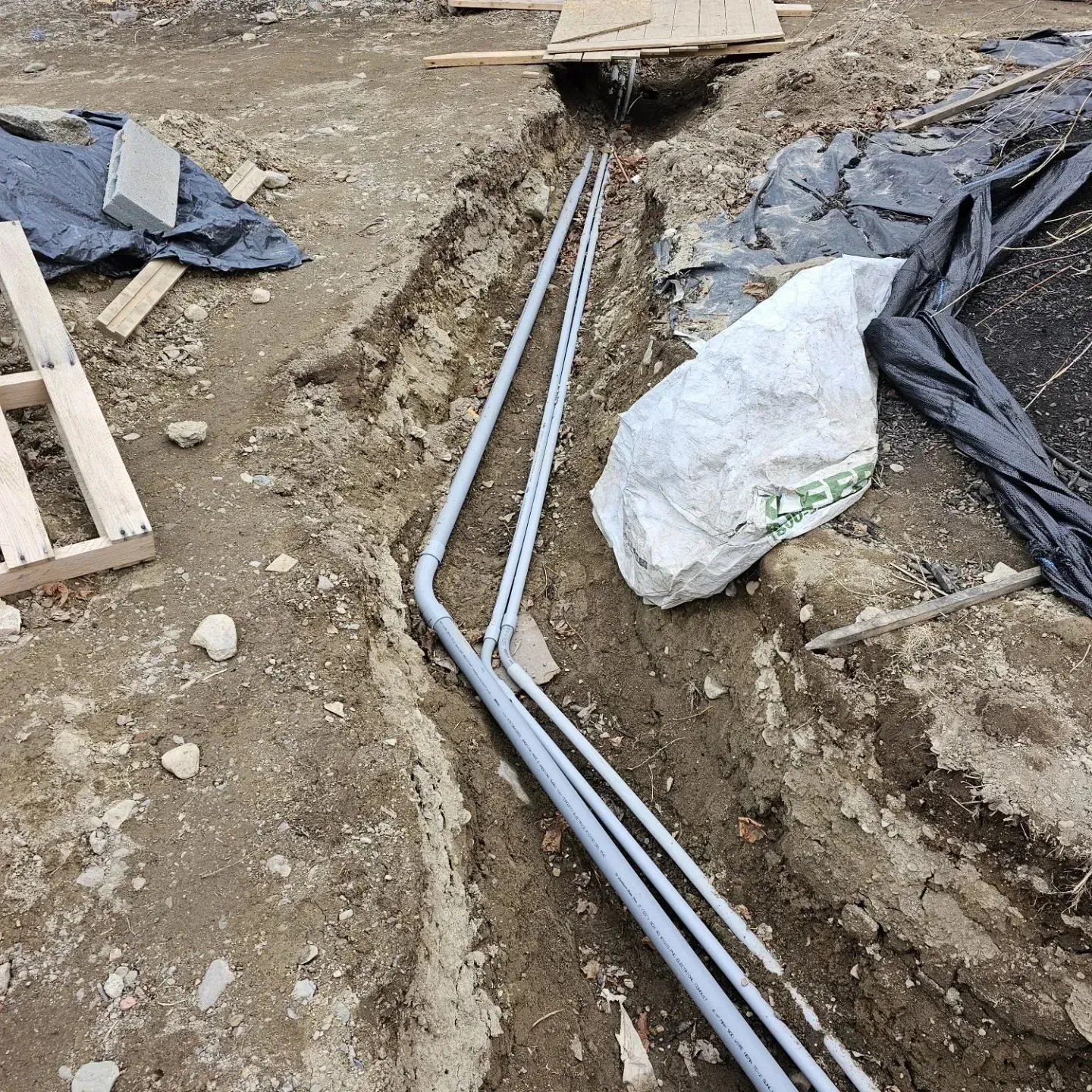 Trenches with gray electrical conduit laid in the dirt at a construction site, with tarps and materials nearby