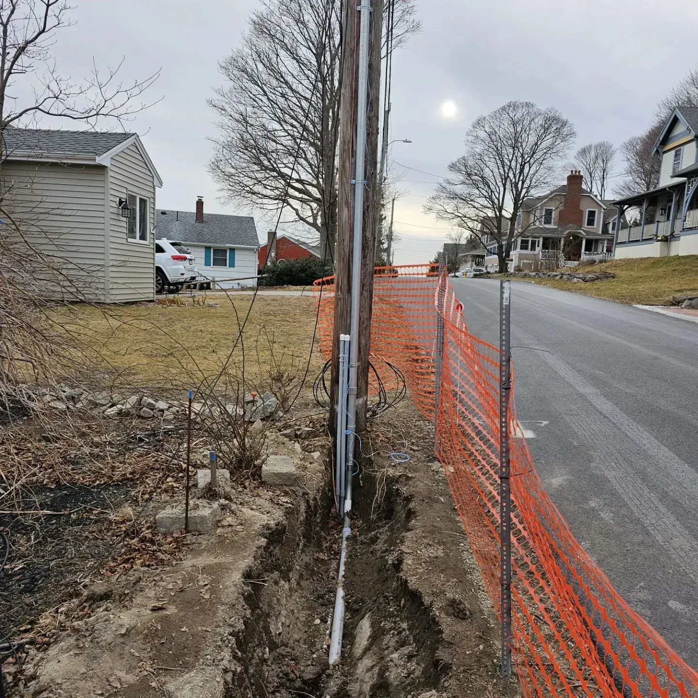 Utility pole in a trench beside a road, with orange safety fencing and houses in the background