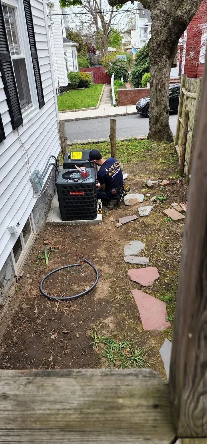 Narrow side yard with a person kneeling beside a black machine near a house wall, with pavers and bare ground.