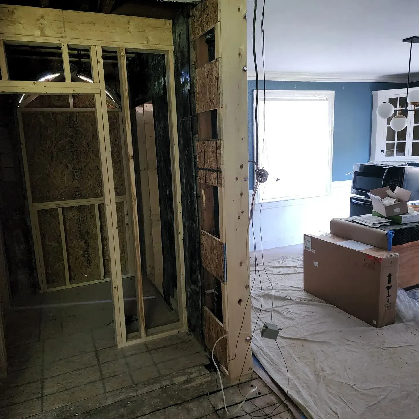 Partially renovated interior: exposed studs and doorway opening to a bright room with furniture and boxes.