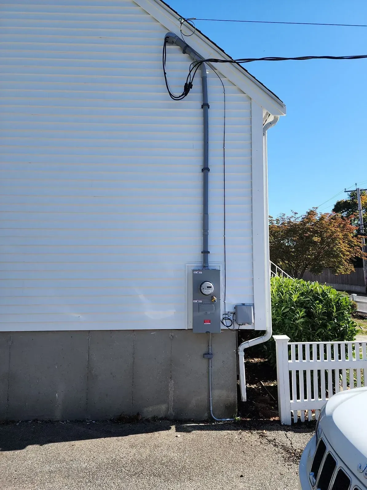 Side of a white house with an electrical meter box and conduit beside a white fence and car hood.