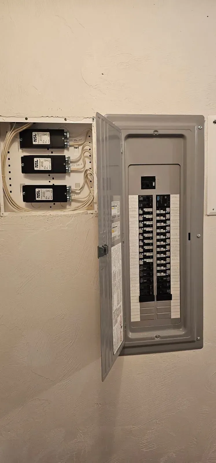 Open electrical panel with breakers beside three wall switches in a beige hallway.