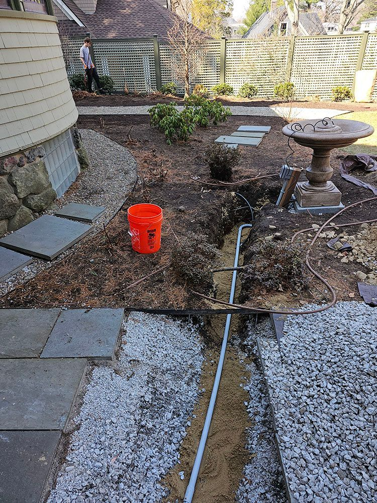 Backyard under construction with a trench, pavers, gravel, and a red bucket beside a birdbath