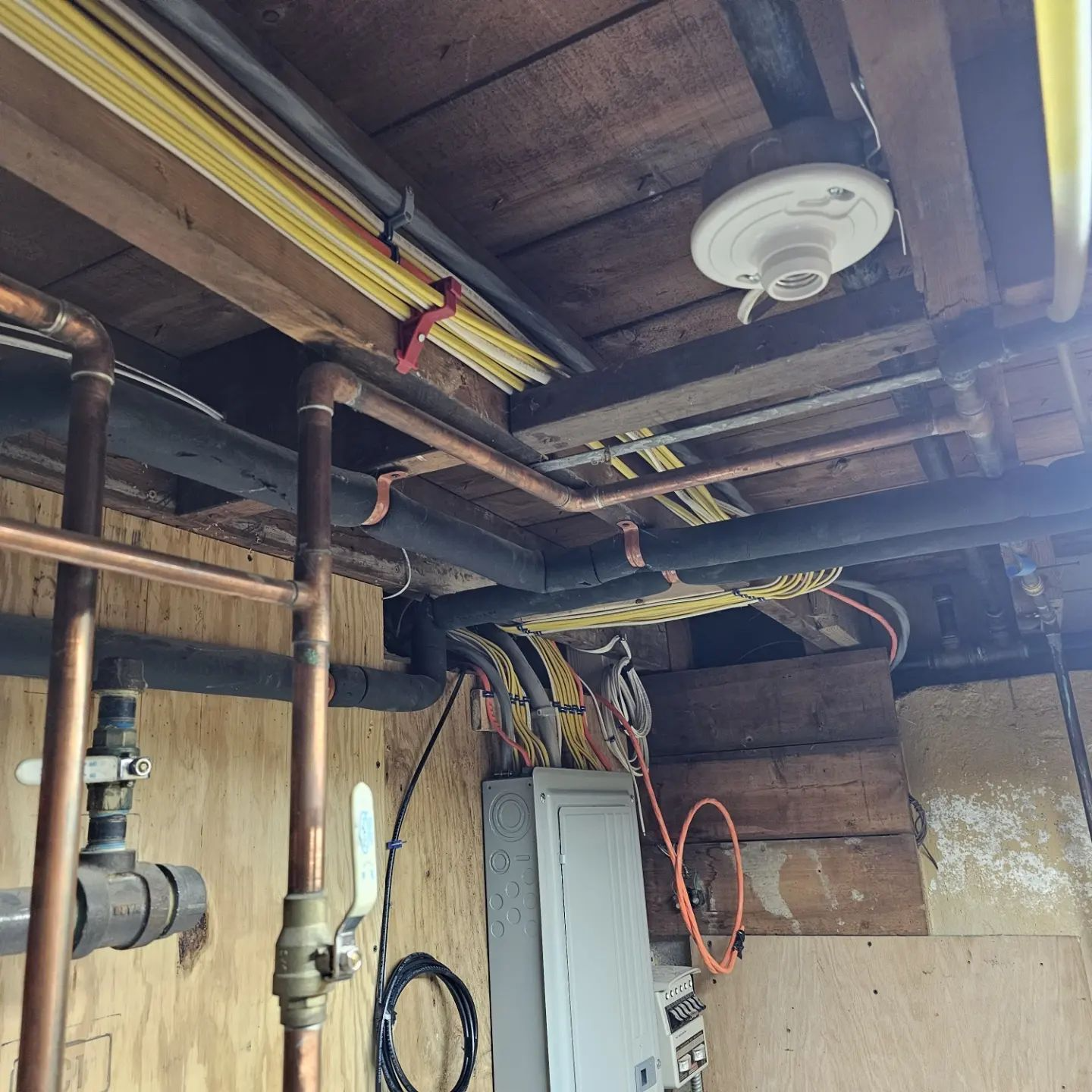 Basement utility room ceiling with exposed pipes, wiring, electrical panel, and a smoke detector
