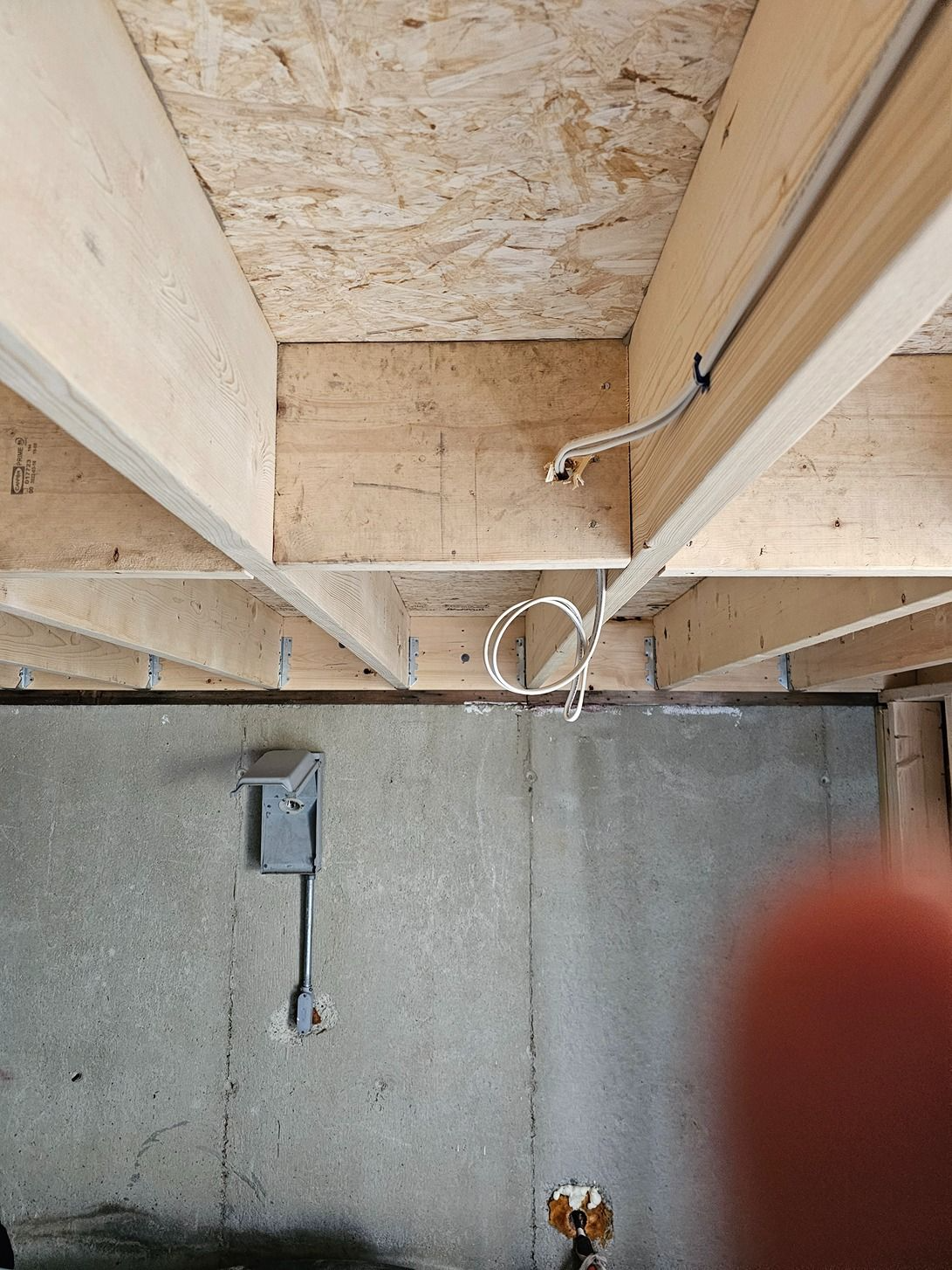 Basement ceiling with exposed wooden joists and wiring above a concrete wall; a finger partly blocks the lower right.