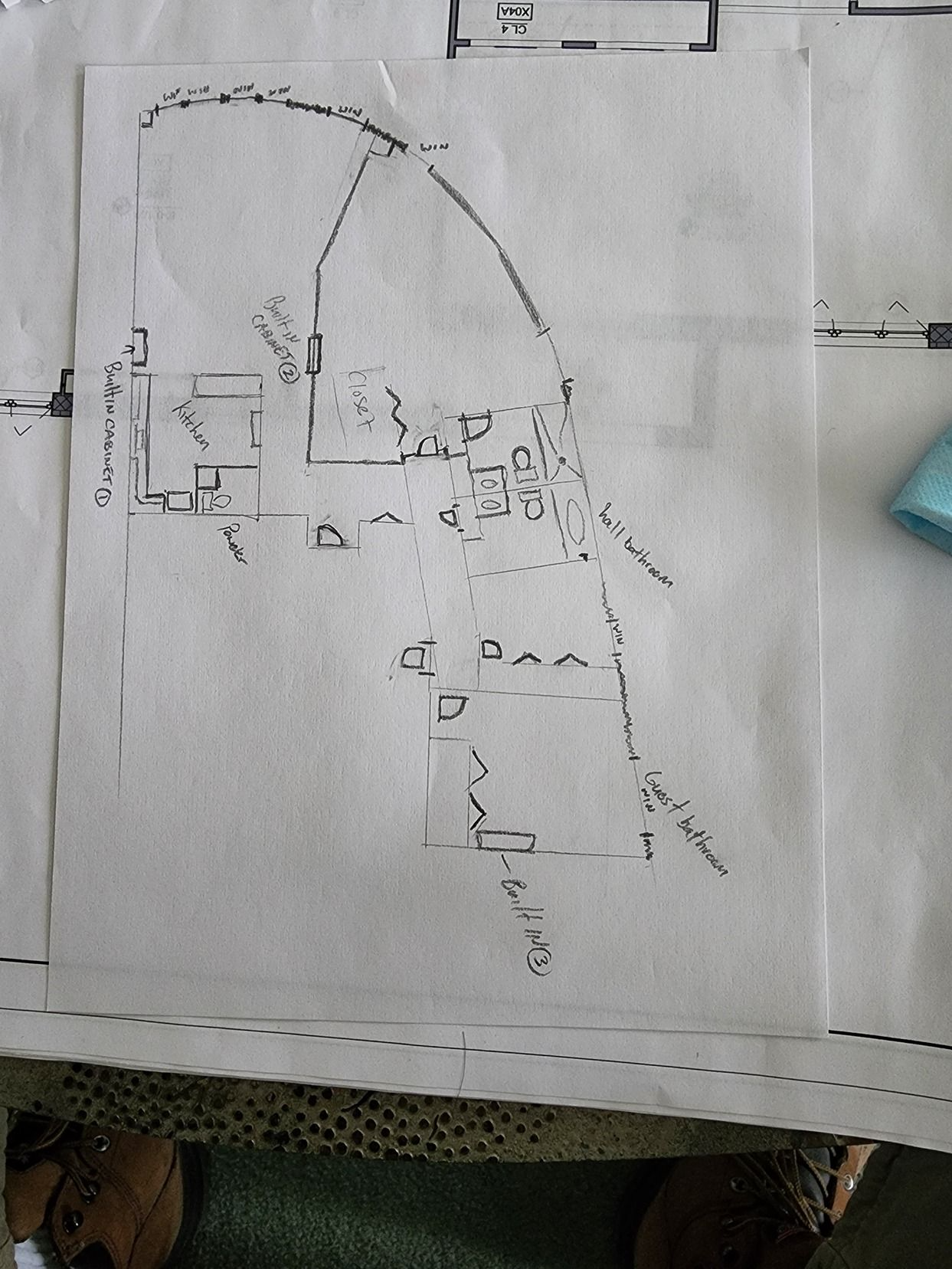 Faint hand-drawn floor plan on white paper, photographed on a table with a blue object at the right edge.
