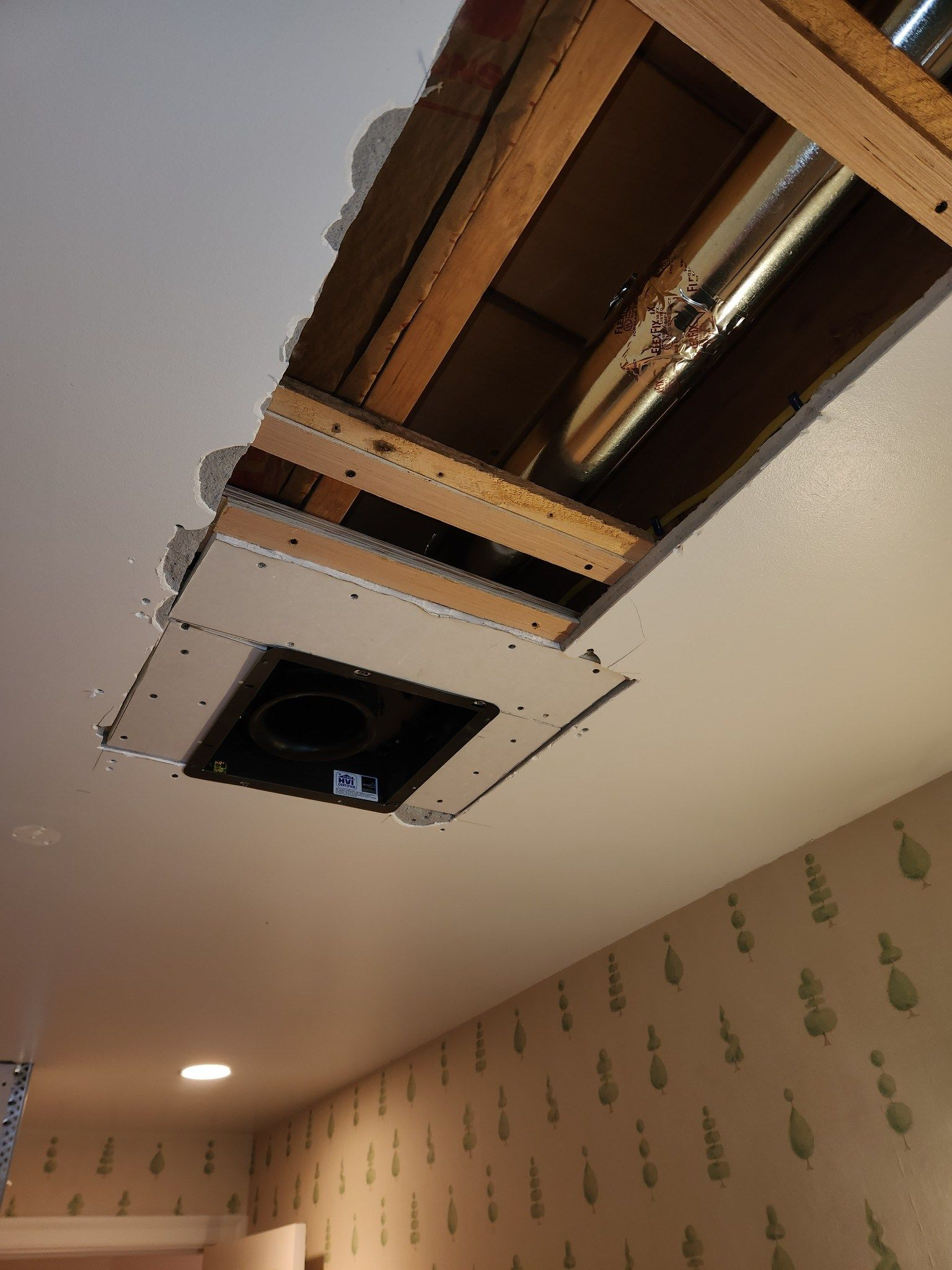 Ceiling vent opening with exposed ductwork and a removed access panel in a room with patterned wallpaper