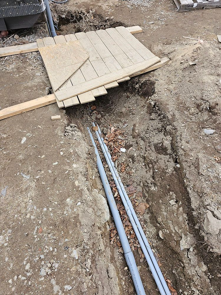 Trench with exposed gray pipes beneath a makeshift wooden plank walkway at a construction site