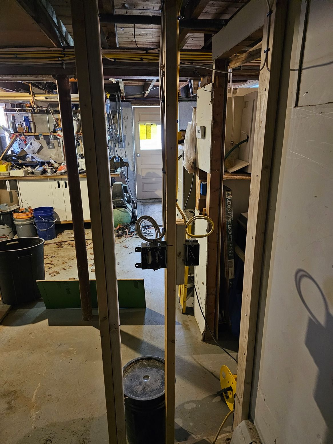 Basement utility area with exposed beams, pipes, shelves, and scattered tools, lit by daylight from a small window