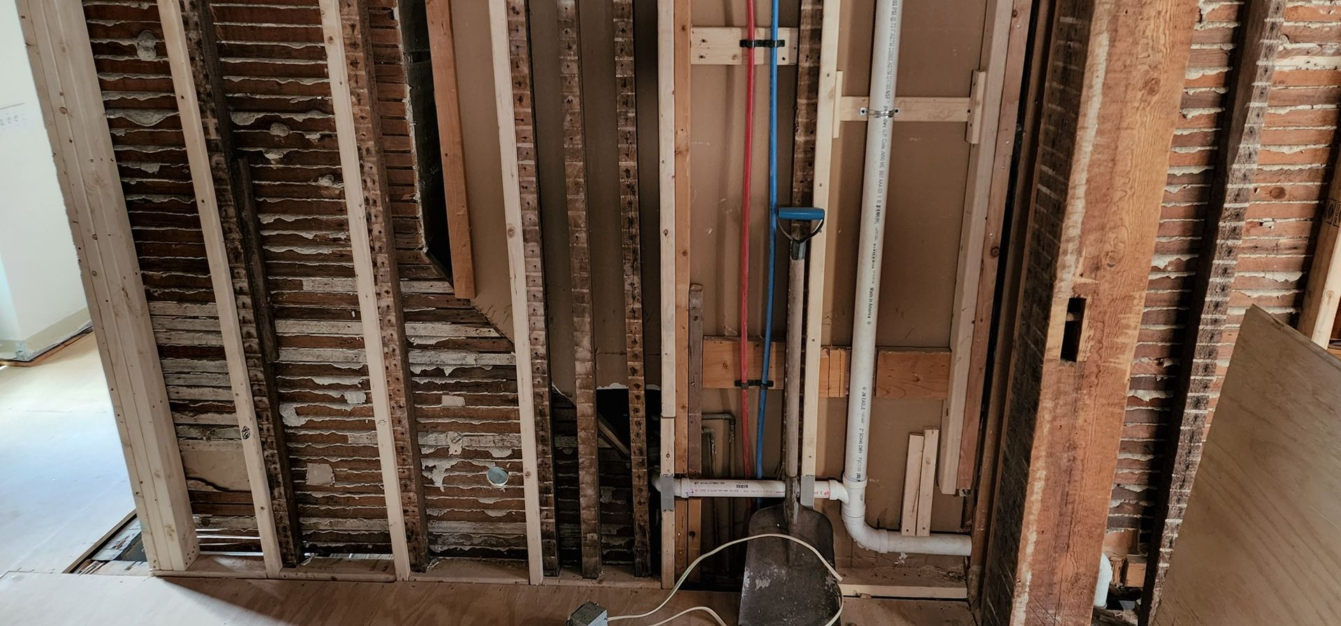 Exposed wall framing and plumbing in a partially renovated interior with wooden studs and debris on the floor