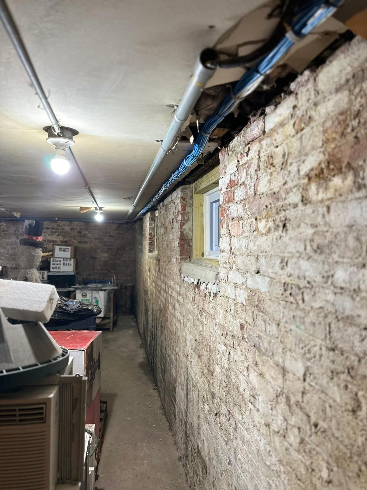 Narrow basement corridor with exposed brick wall, overhead pipes, and a small window, cluttered with equipment on the left.