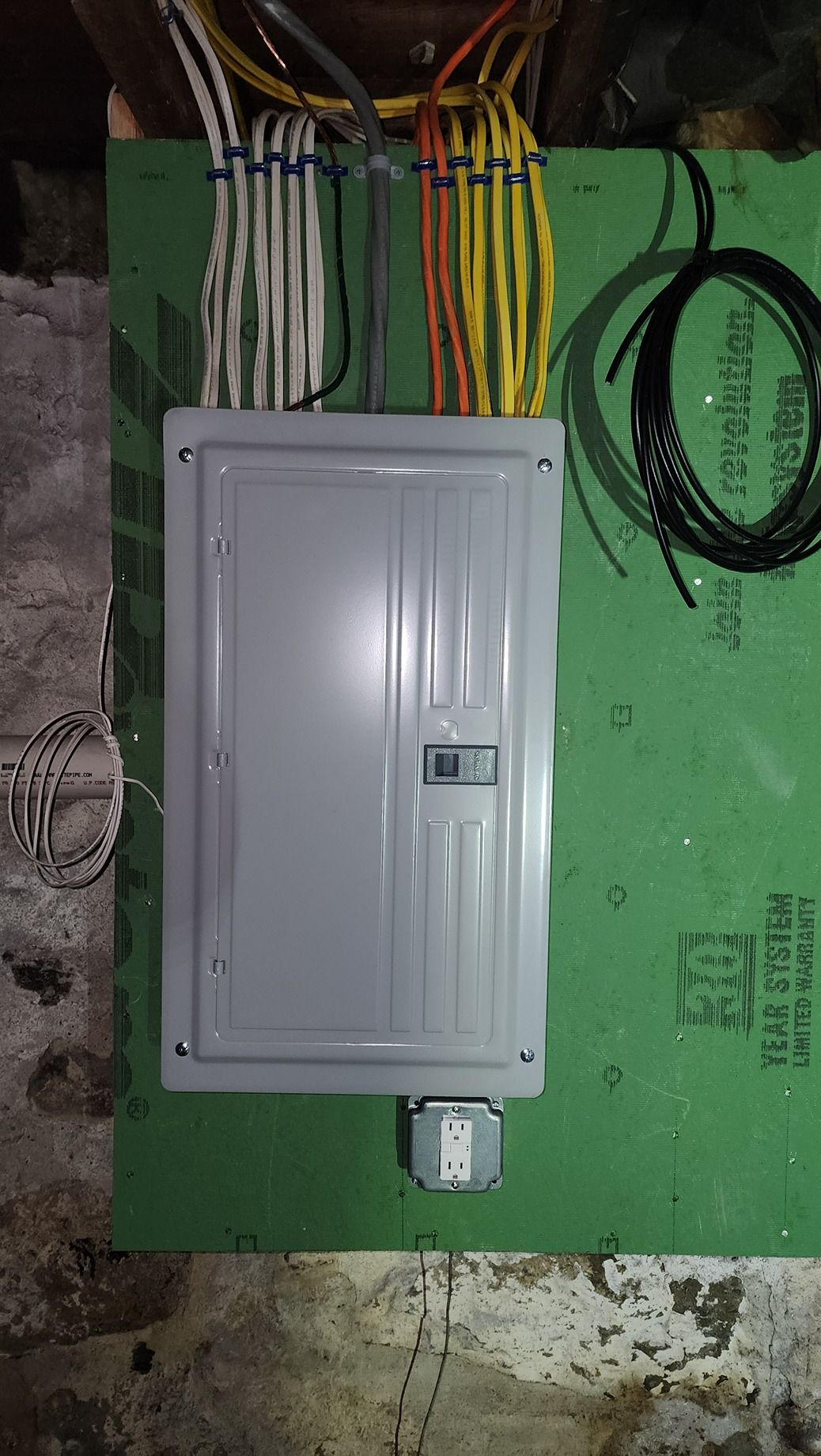 Gray electrical breaker panel on a green wall with exposed colored wires running into the top.
