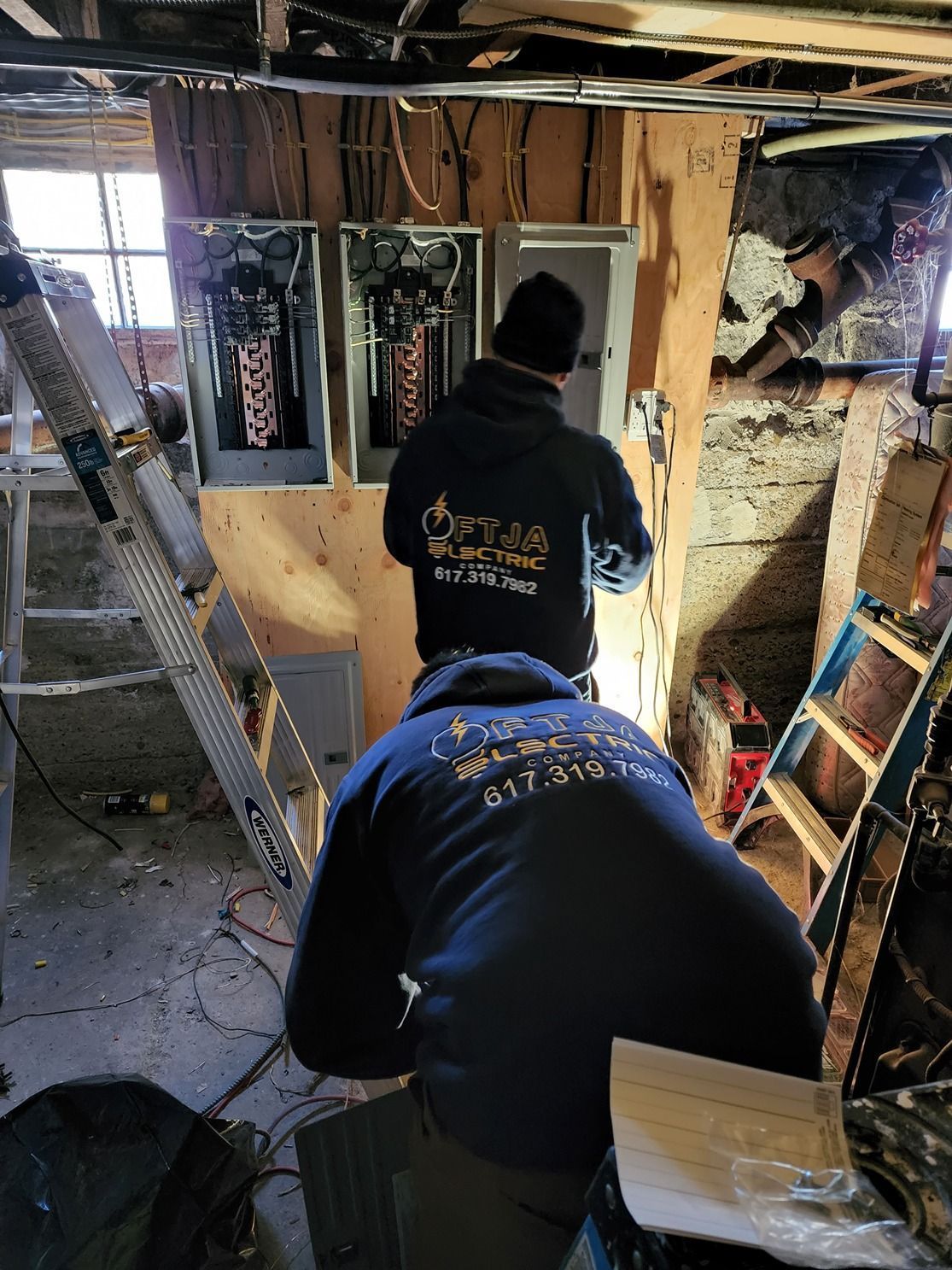 Two workers in hoodies framing an interior wall in a dim, unfinished room with ladders and tools.