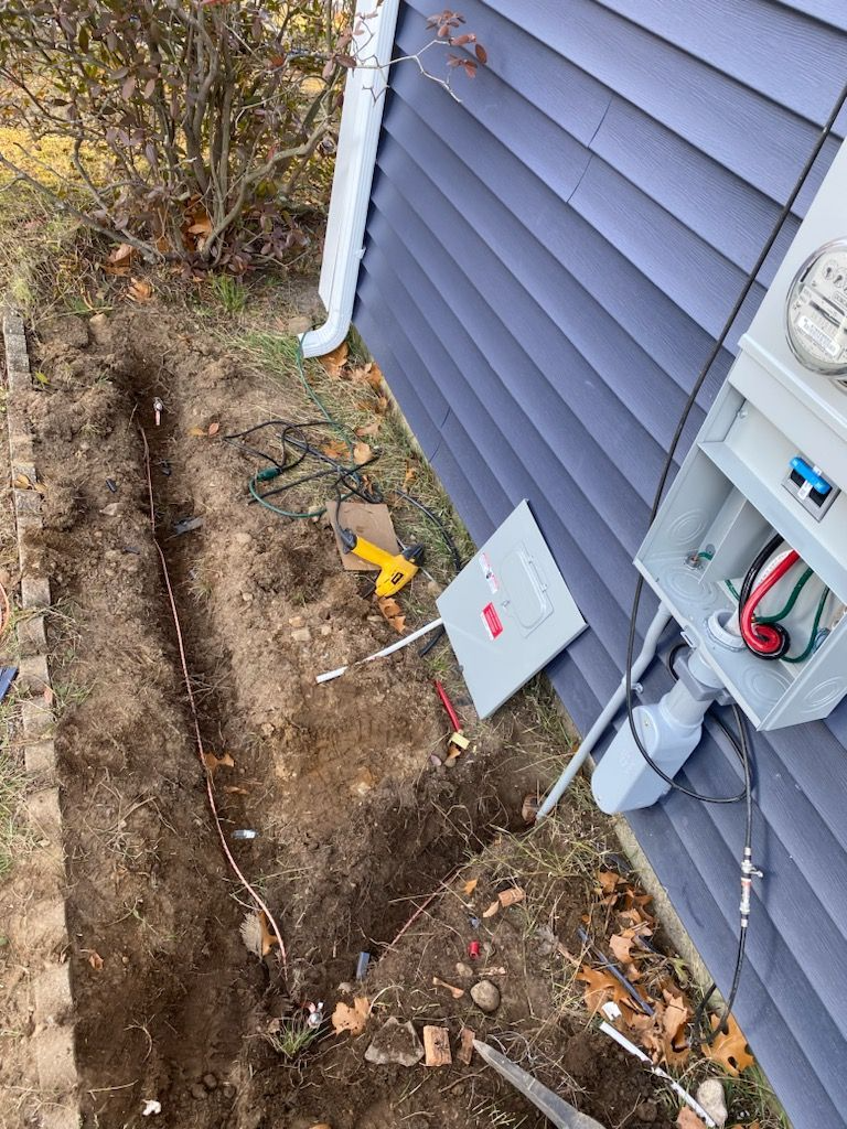 Utility boxes and cables beside a blue house, with a dug trench along the foundation.