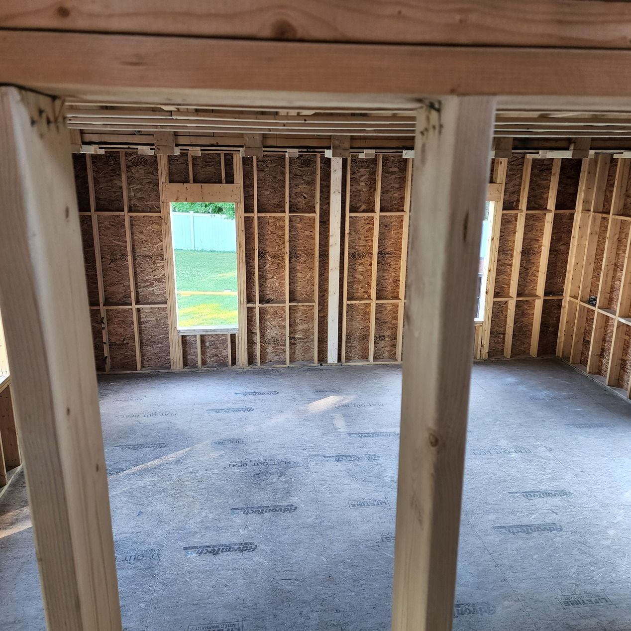 Interior of an unfinished room with exposed framing, subfloor, and a small window showing green grass outside.