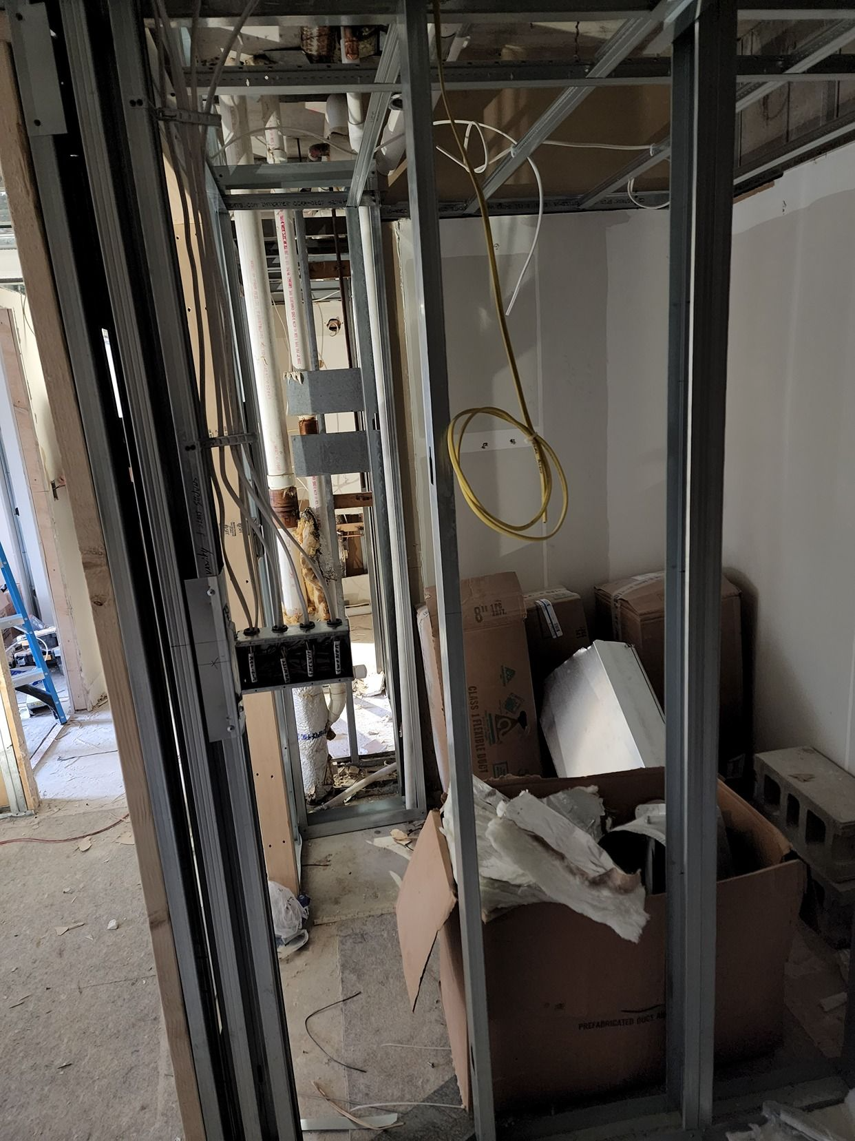 Narrow construction corridor with exposed framing, hanging wires, debris, and a cardboard box on the floor