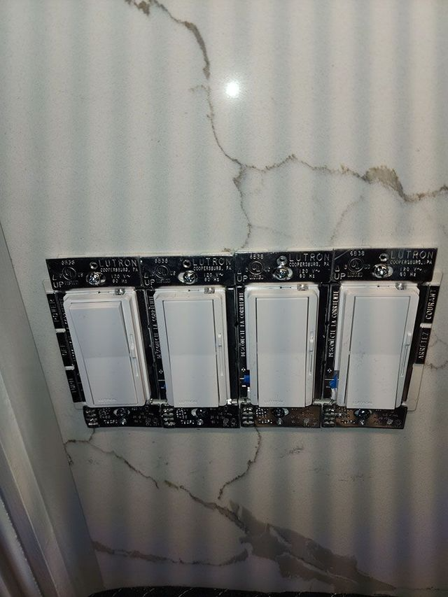 Four white wall light switches on a black mounting plate against a gray marbled wall.
