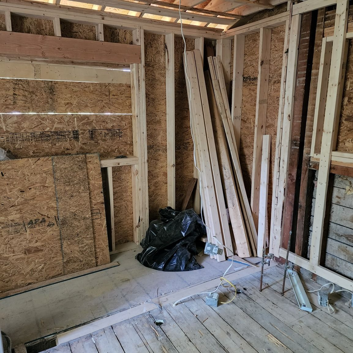 Unfinished room with exposed framing, OSB walls, lumber boards, and a black trash bag on the floor.