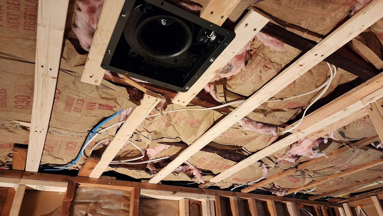 Exposed attic ceiling framing with insulation and a black ventilation fan installed between joists