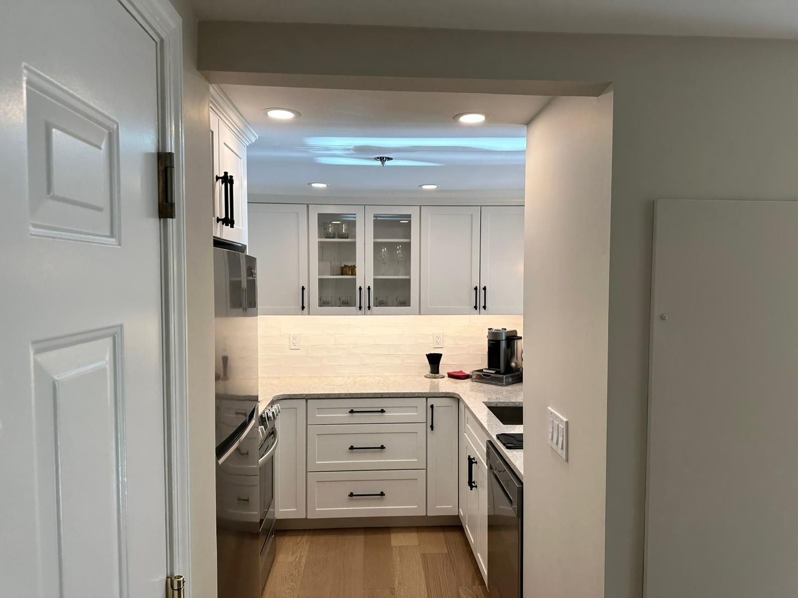 Modern white kitchen seen through an open doorway, with cabinets, stainless appliances, and warm under-cabinet lighting