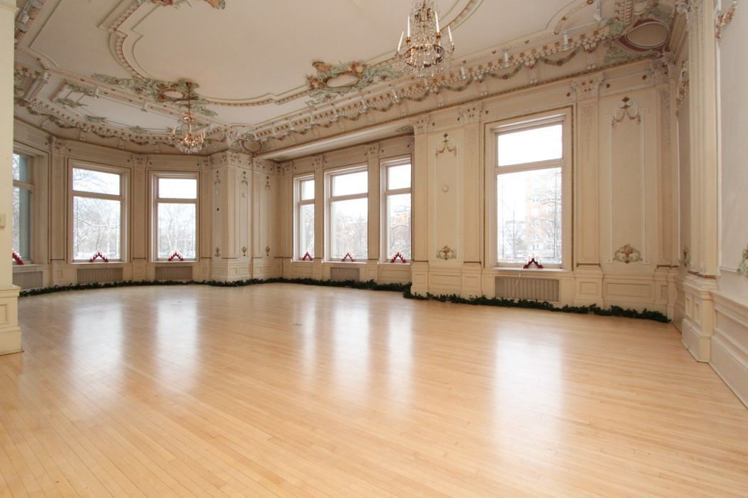 Empty, ornate ballroom with light wood floor, large windows, and decorative ceiling molding.