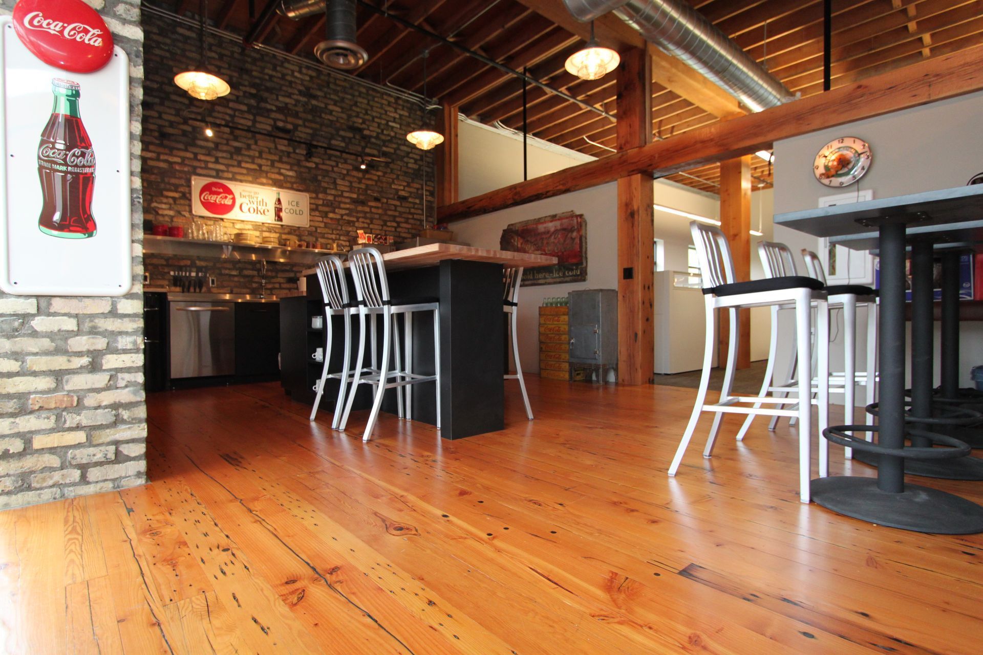Interior with wooden floor, brick wall, bar with stools, Coca-Cola signs, and overhead lighting.