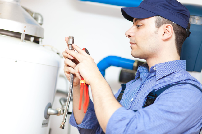 Plumber in blue uniform fixing water heater with pliers in an indoor setting.
