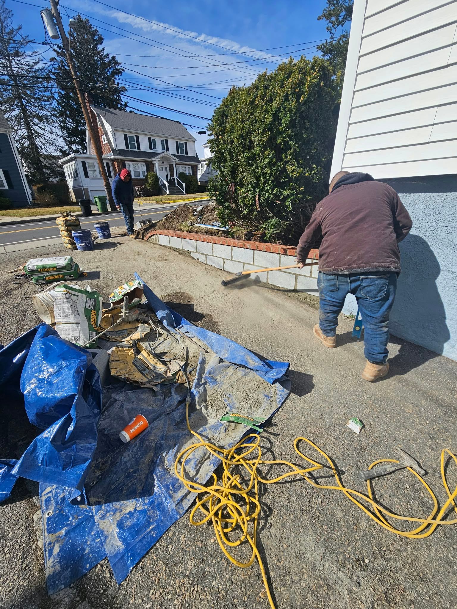 Worker laying pavers along a sidewalk beside a house, with tools, tarps, and extension cords nearby.