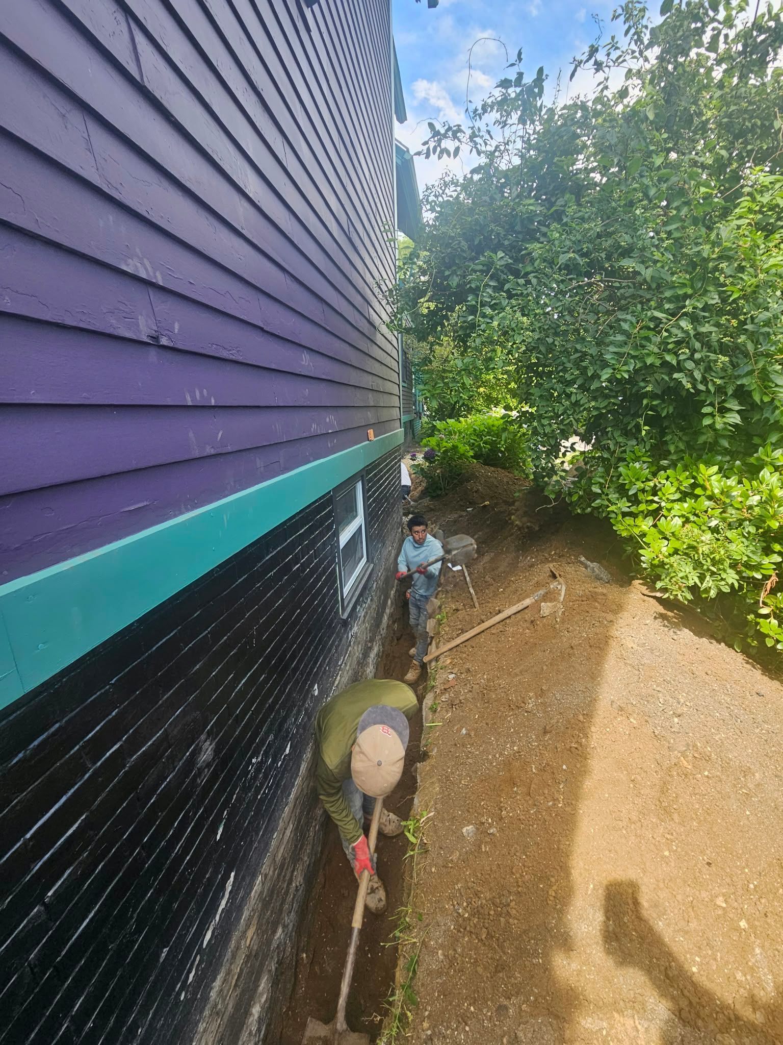 Side of purple house with teal trim beside a dirt path and green trees in bright sunlight