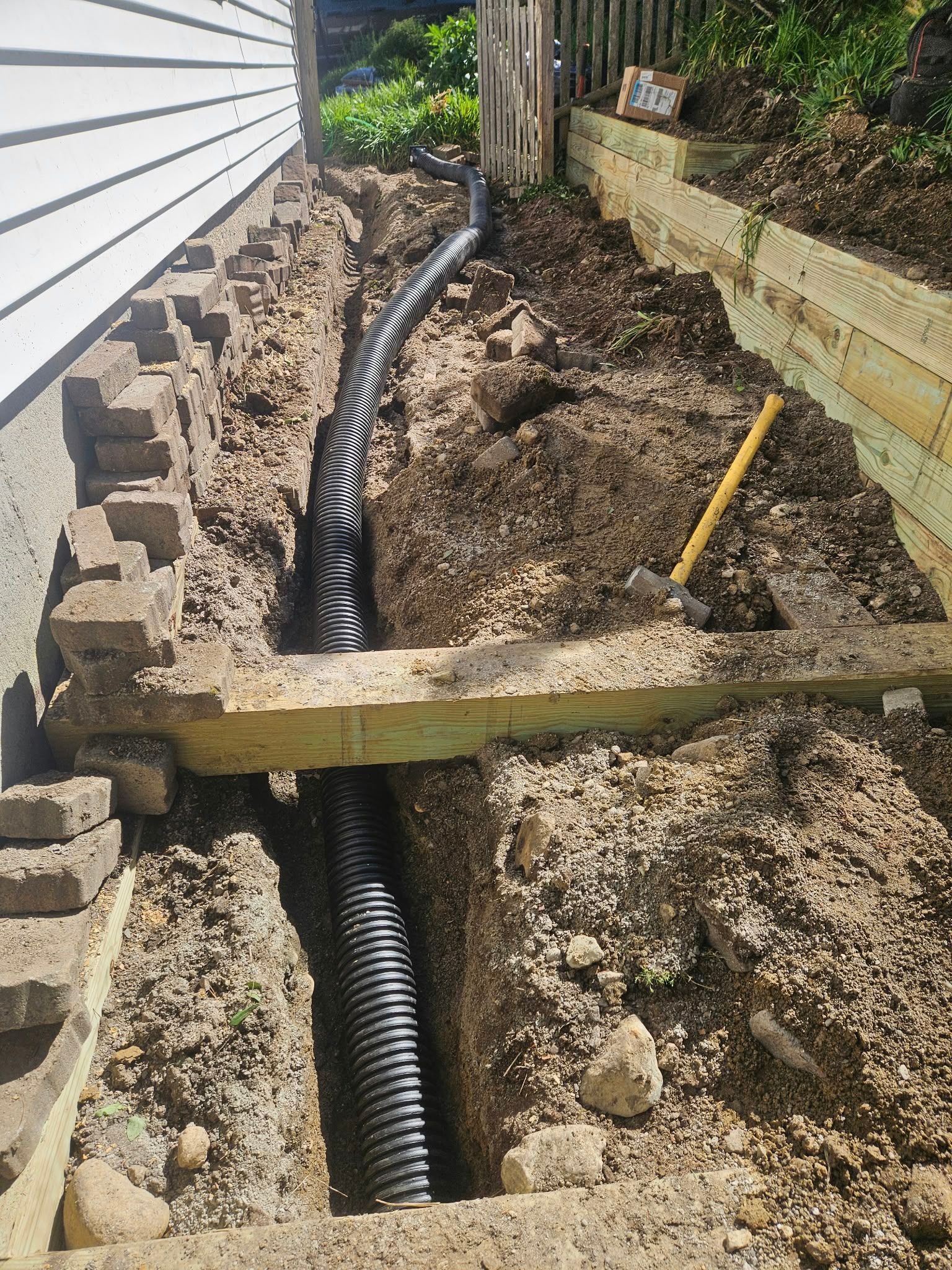 Construction trench with black drainage pipe and soil beside a house foundation.