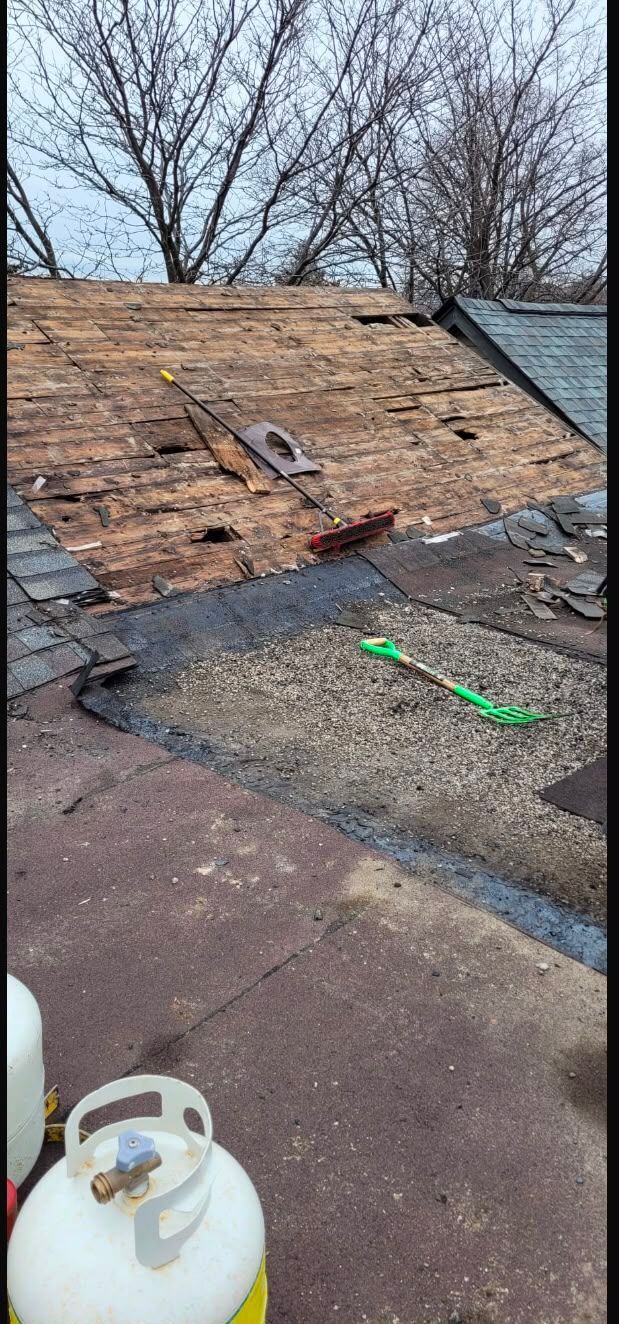 Damaged roof with exposed plywood, scattered debris, and a propane tank on a flat rooftop.