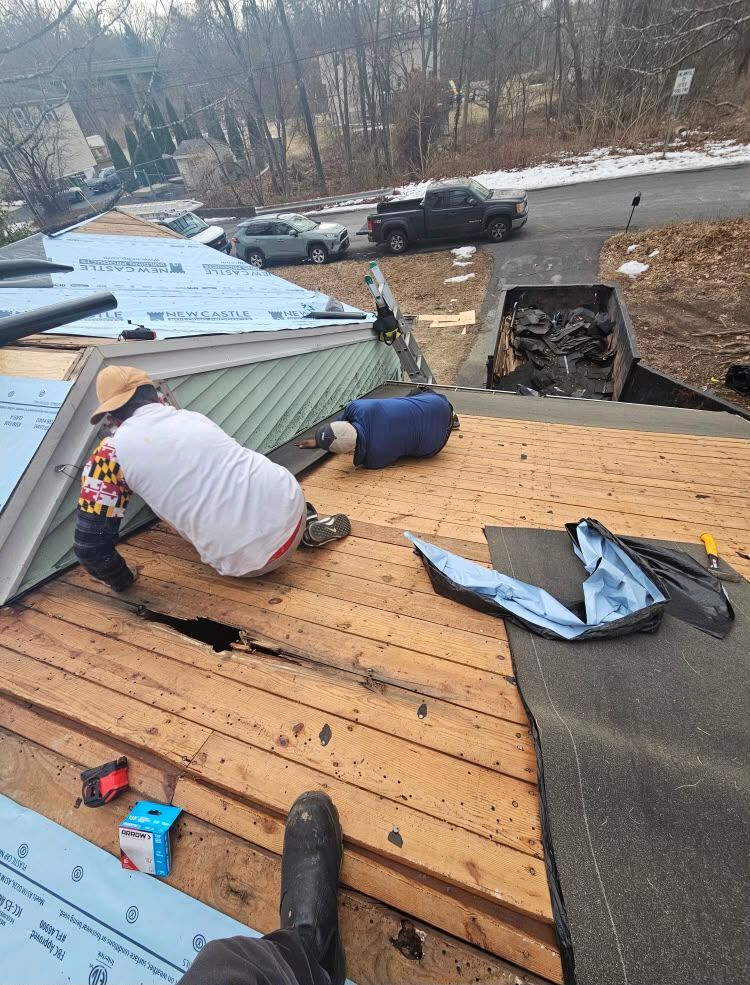 Worker kneeling on a roof, installing plywood and underlayment near damaged shingles and a chimney.