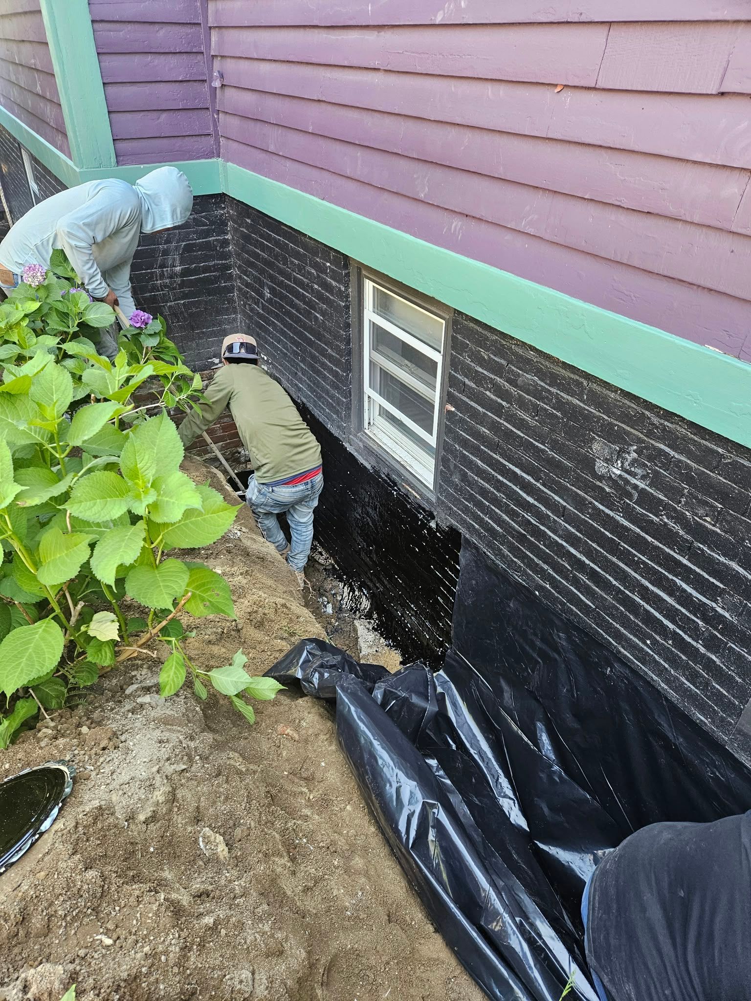 Worker in white protective gear inspecting a house foundation beside black waterproofing and soil.