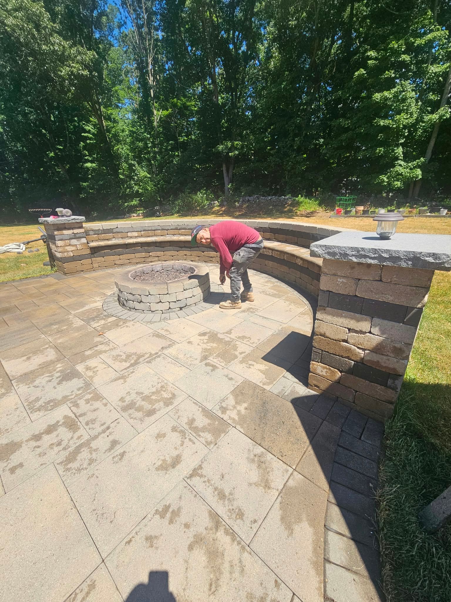 Person in a red shirt bending over a stone fire pit in a curved outdoor patio surrounded by trees
