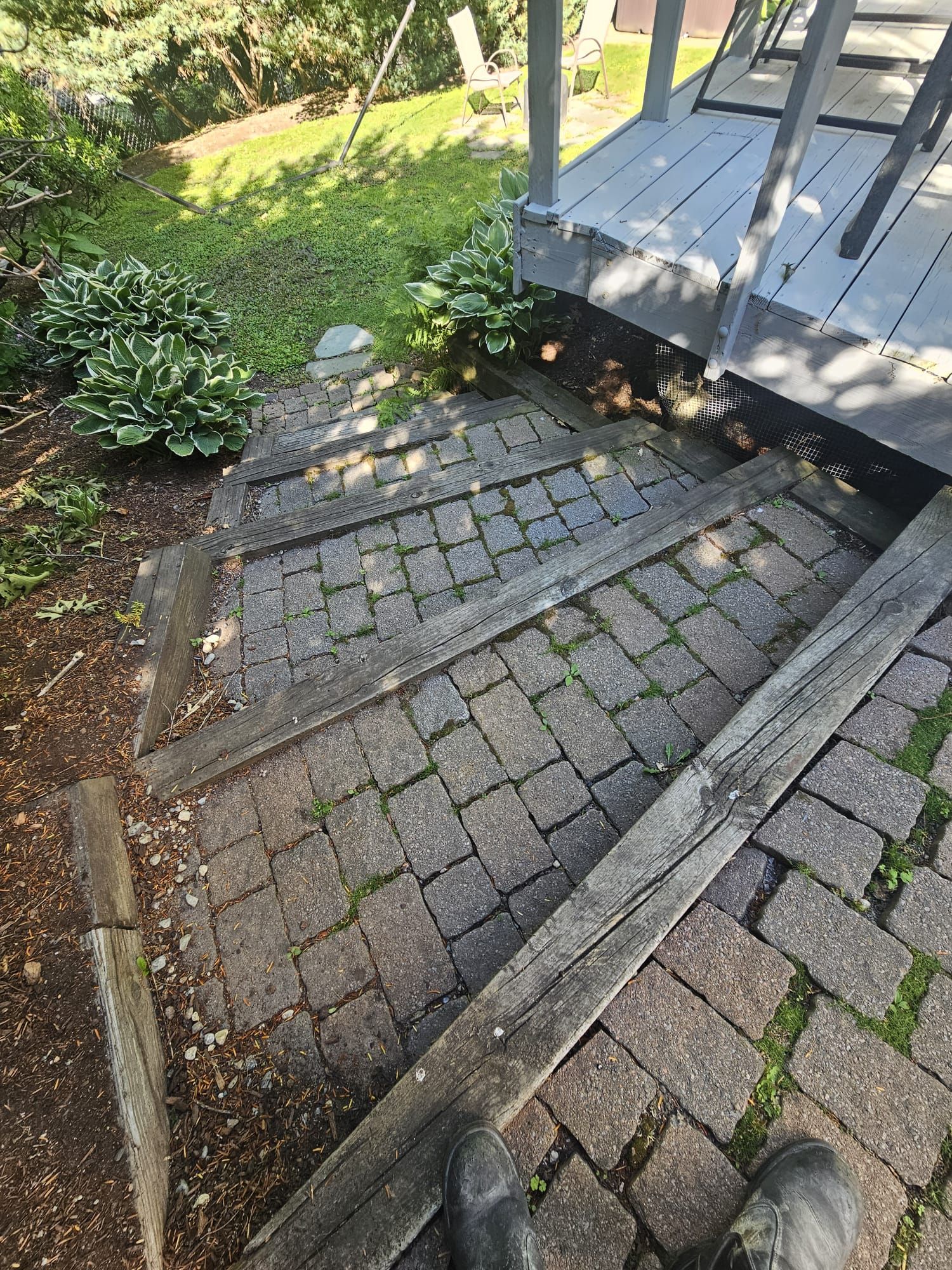 Broken brick patio beside a wooden deck, with uneven, sunken pavers and a grassy yard nearby