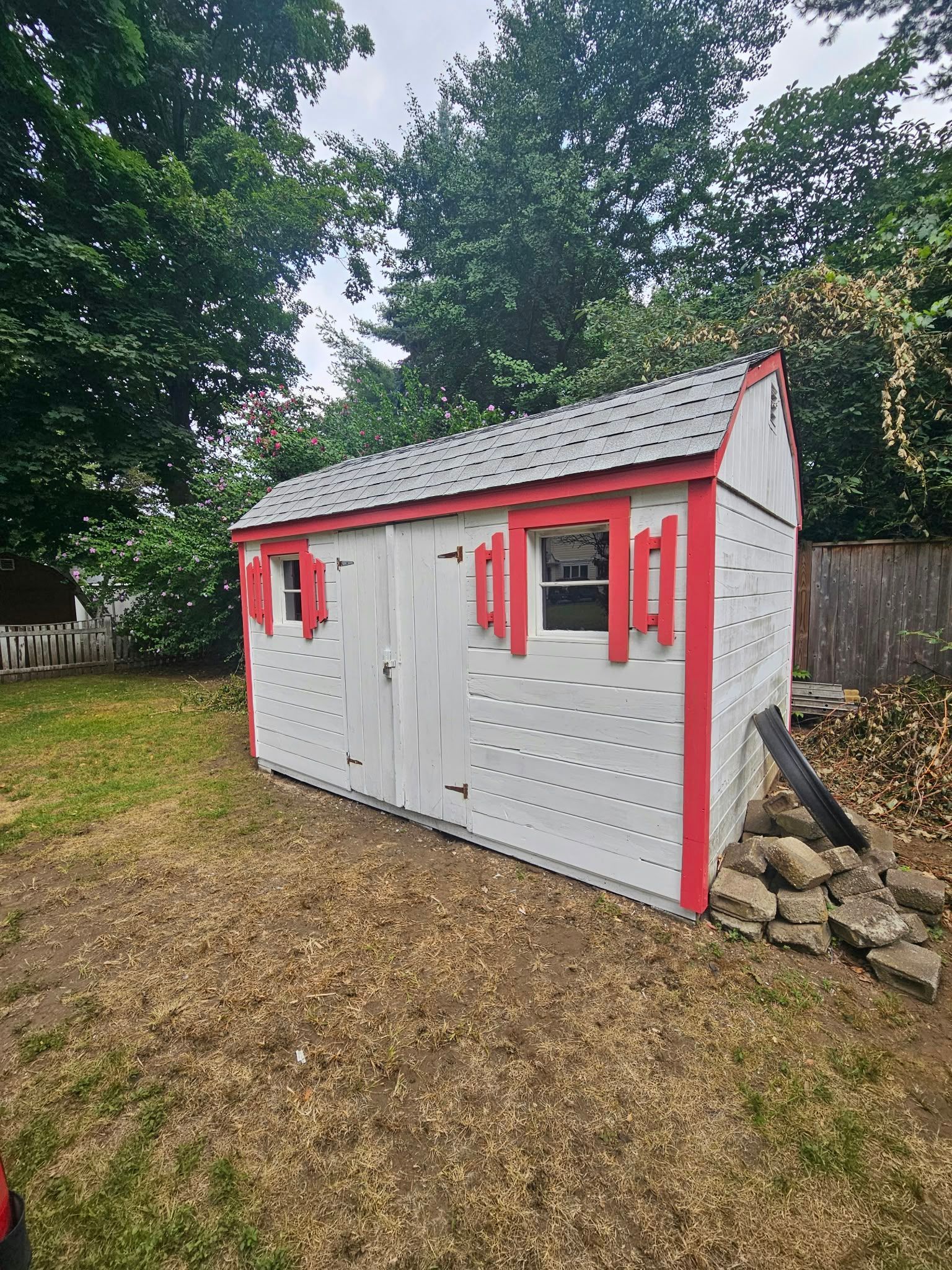 Small white shed with red trim and windows in a grassy yard, surrounded by trees and a wood fence.