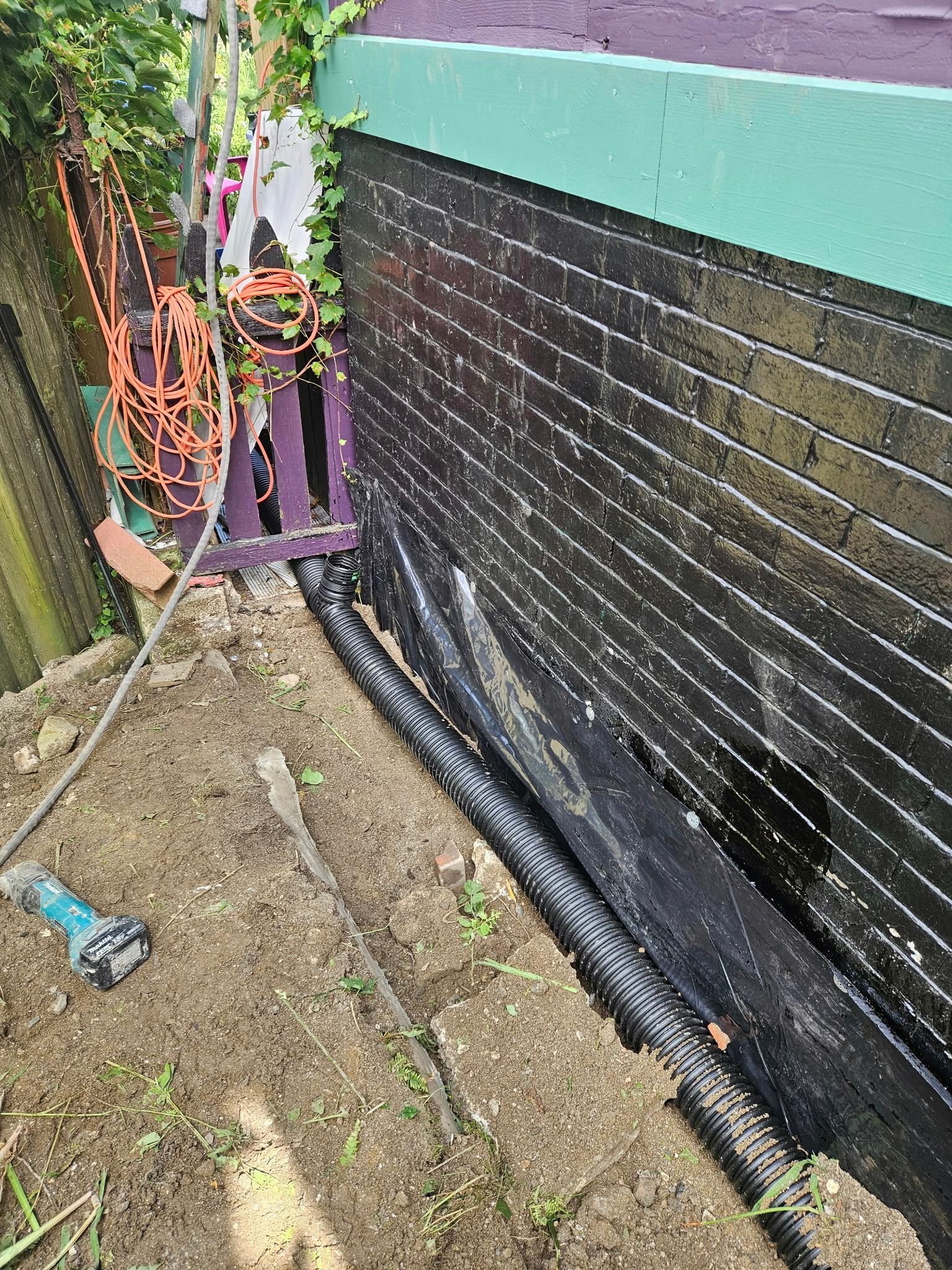 Black brick wall with green trim beside a dirt path and orange/purple garden furniture on the left