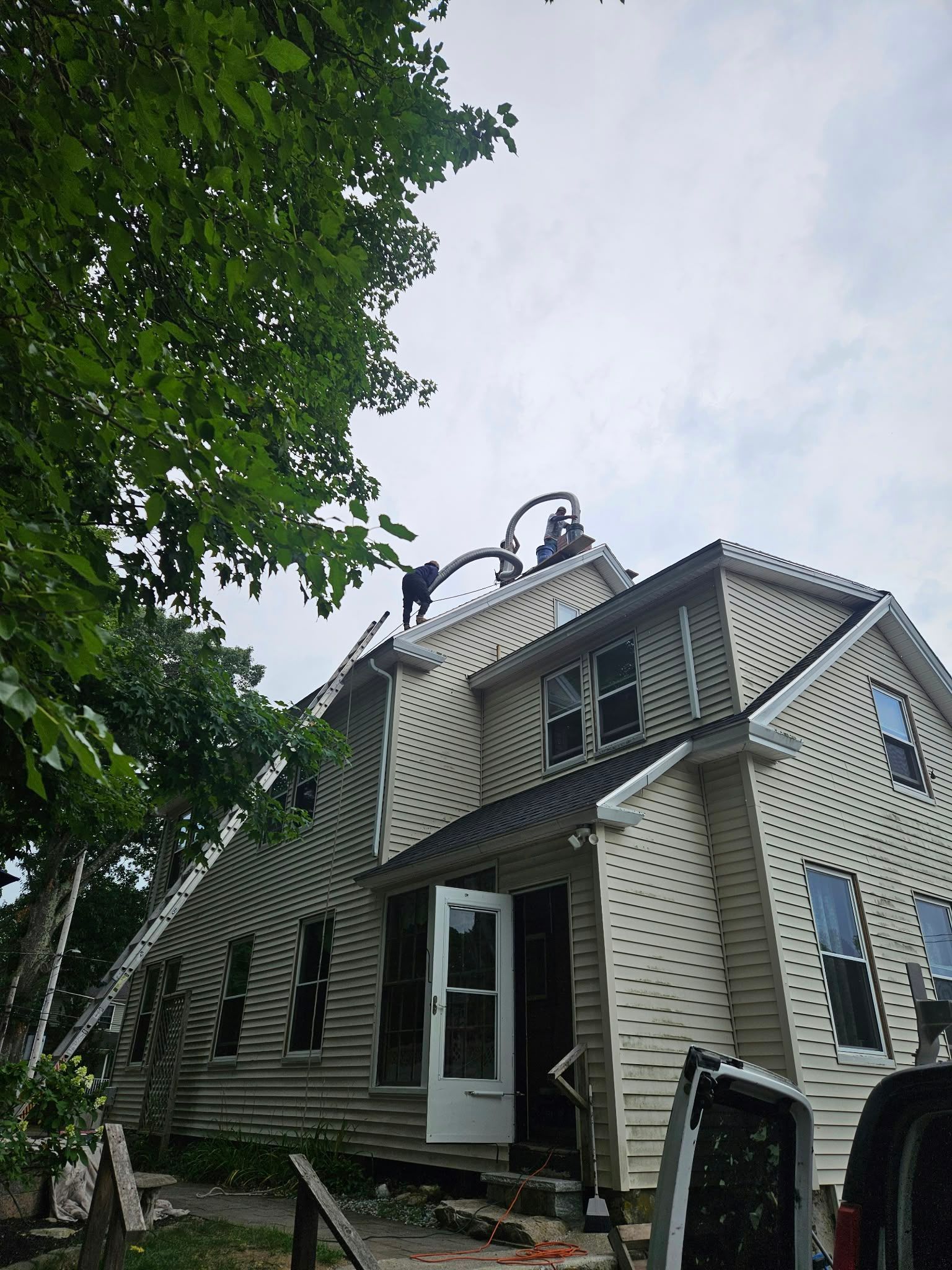 Two-story house with striped siding and open doorway, viewed from a leafy yard under cloudy sky