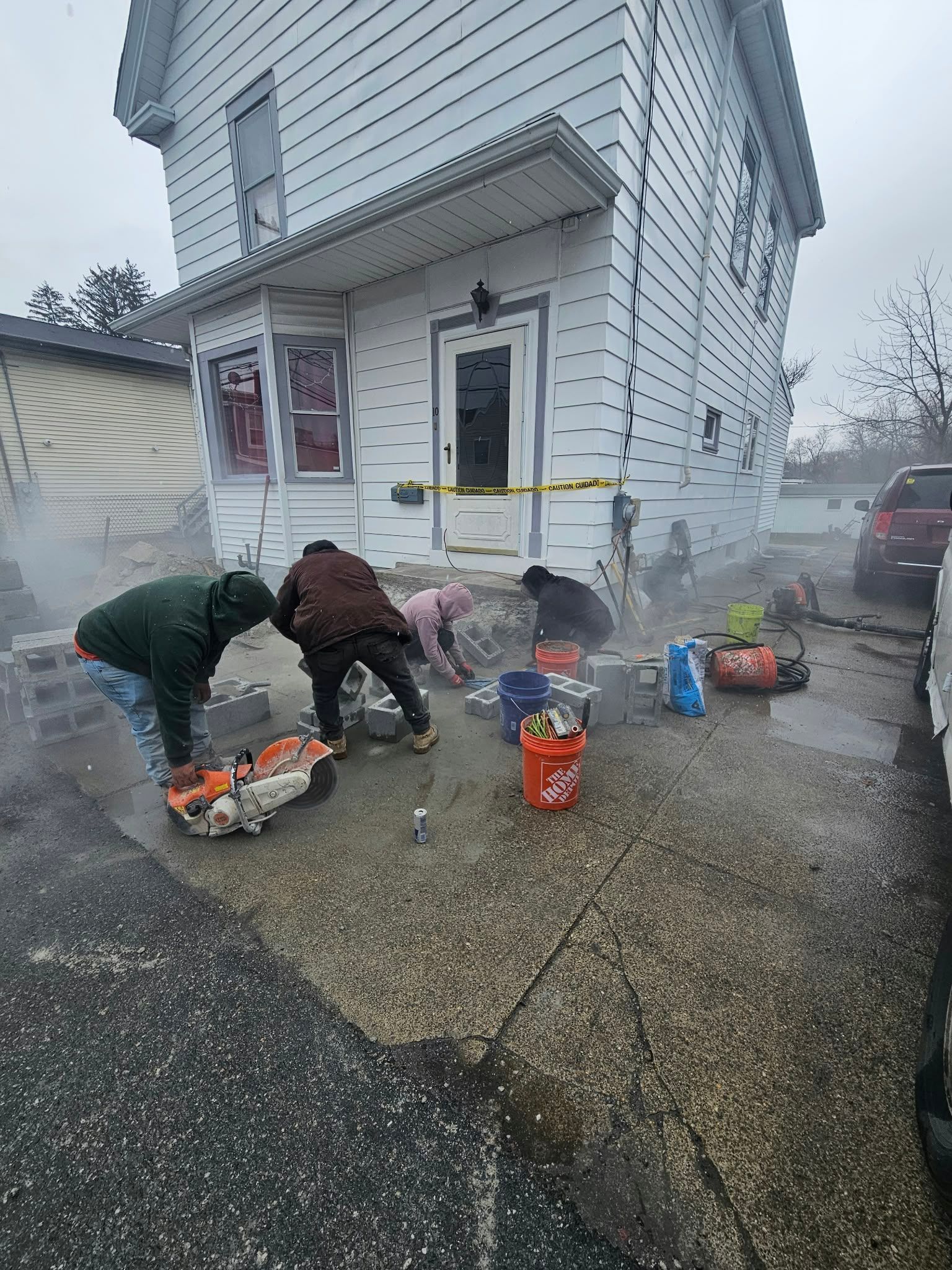 Two people crouch outside a white house, handling supplies and buckets on a wet driveway in cold, rainy weather