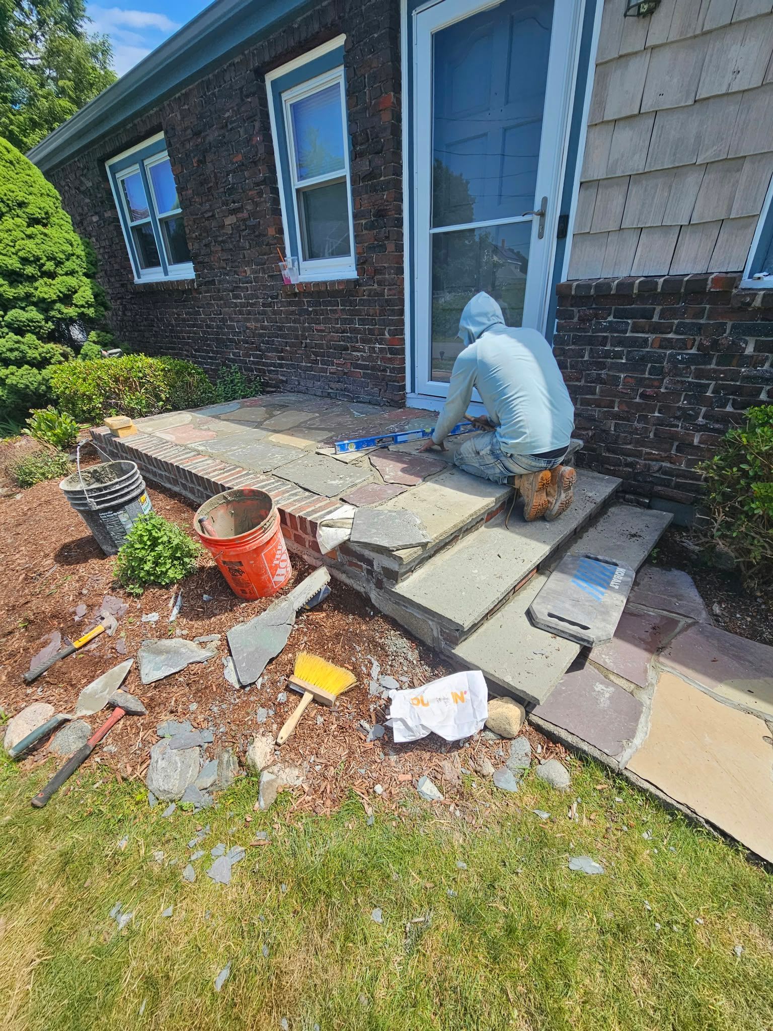 Worker repairing front steps beside a brick house, with tools and materials scattered around.