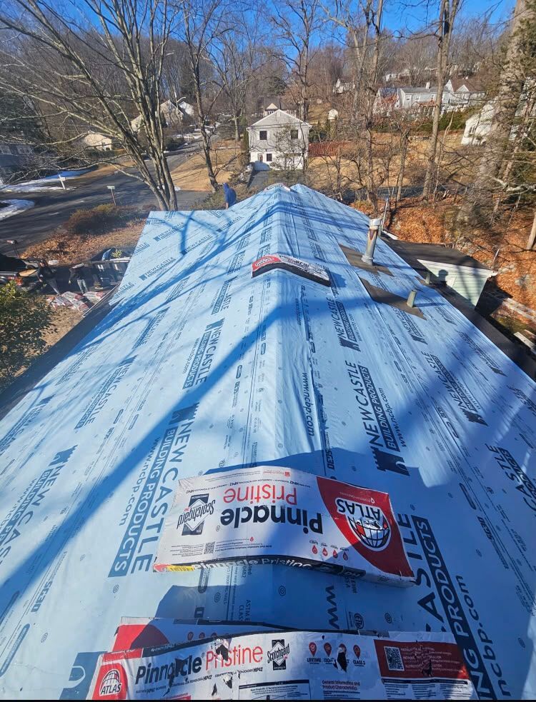 Blue roof underlayment on a house under construction, with trees and a neighboring home in the background.