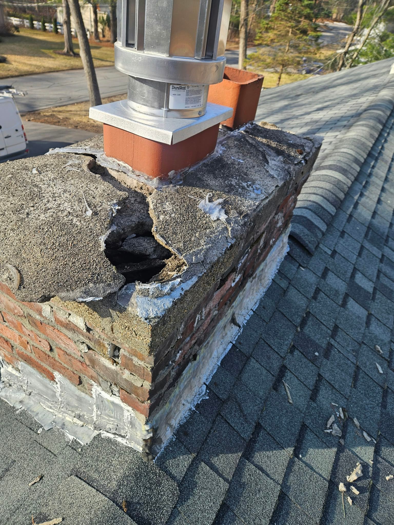 Rooftop chimney base with crumbling mortar and a hole in the brickwork beside a paved walkway.