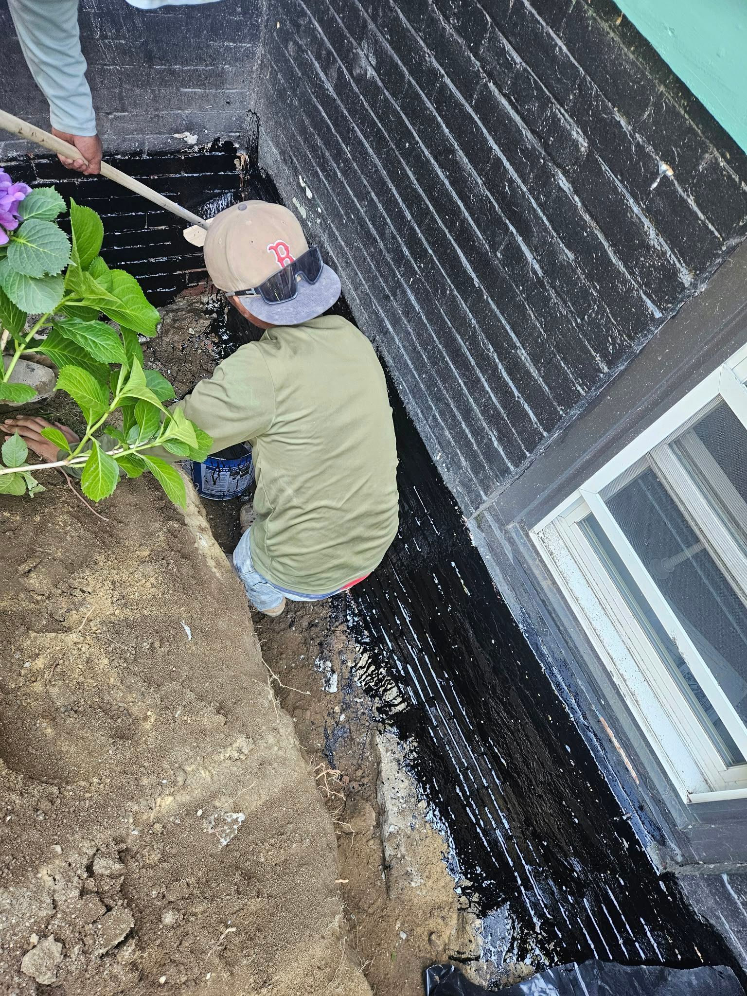 Worker digging along a brick house foundation beside a window, with soil piled nearby.